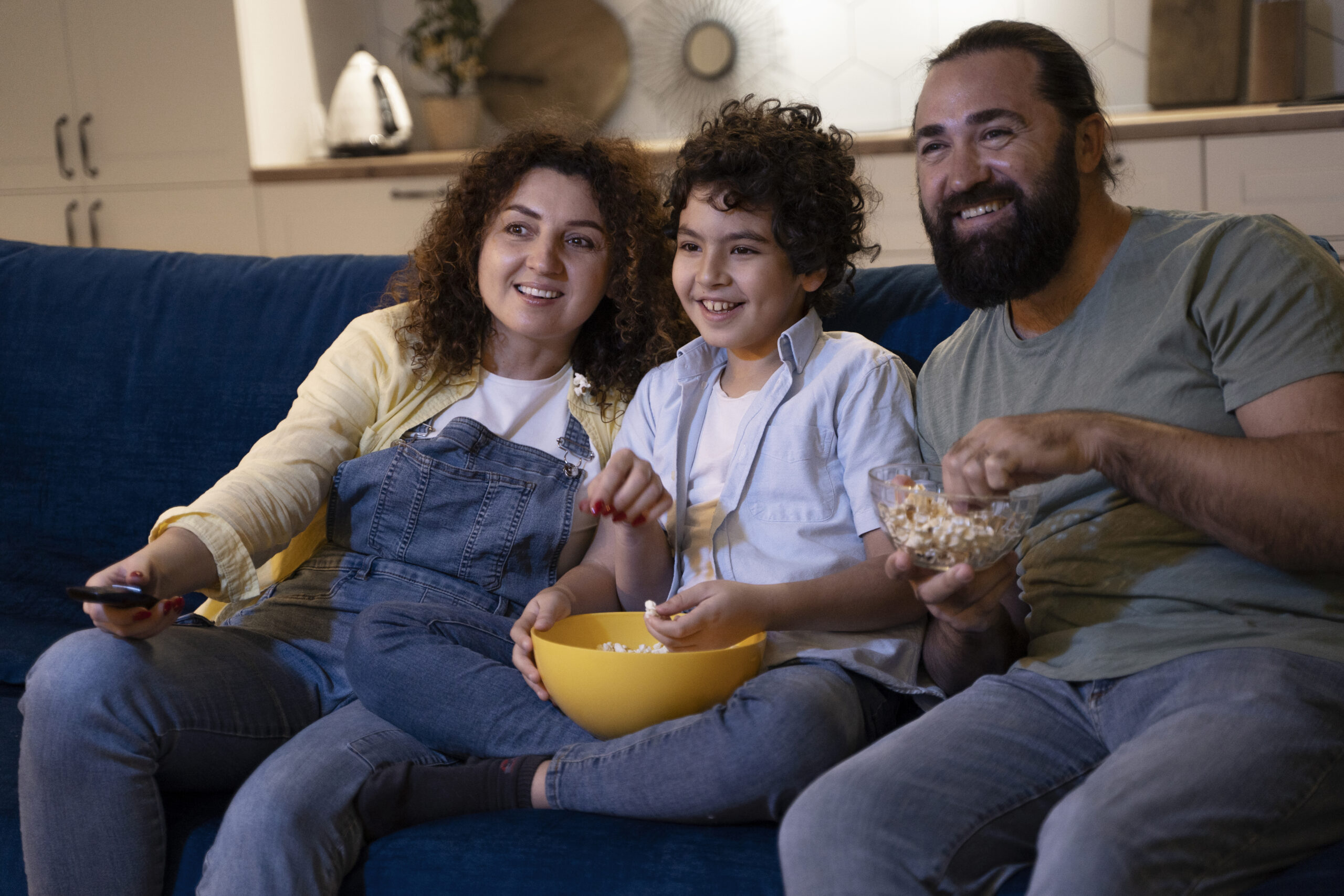 Feature image Família sorridente sentada no sofá assistindo a um filme, comendo pipoca e aproveitando um momento de lazer em casa durante as férias escolares.