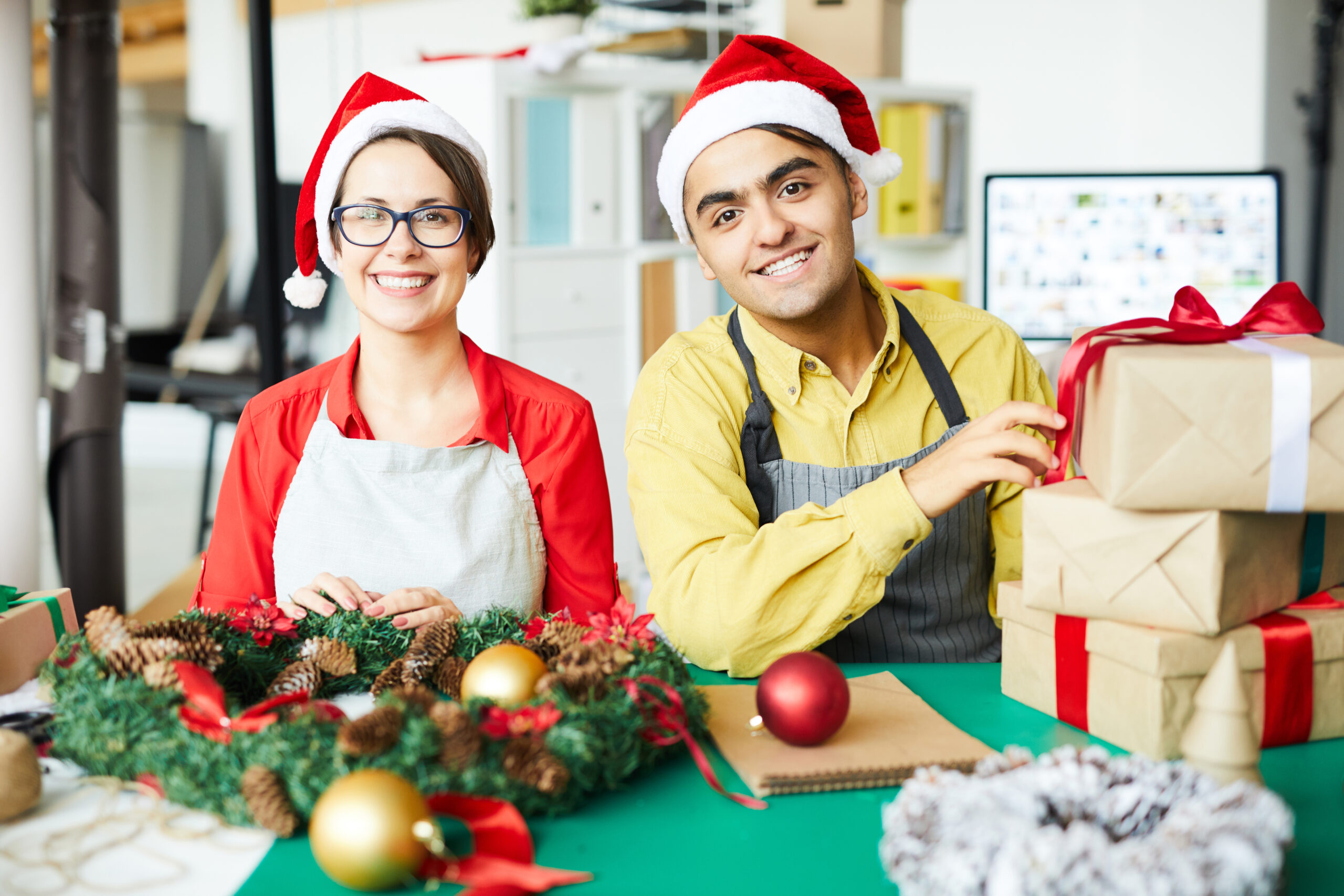 Feature image Dois jovens colegas de trabalho sorriem para a câmera usando gorros de Papai Noel. A mulher, à esquerda, usa óculos, uma camisa vermelha e um avental branco, posicionando as mãos sobre uma guirlanda de Natal feita de pinhas. O homem, à direita, veste uma camisa amarela e um avental cinza, tocando em uma pilha de presentes embrulhados em papel pardo com fitas vermelhas.