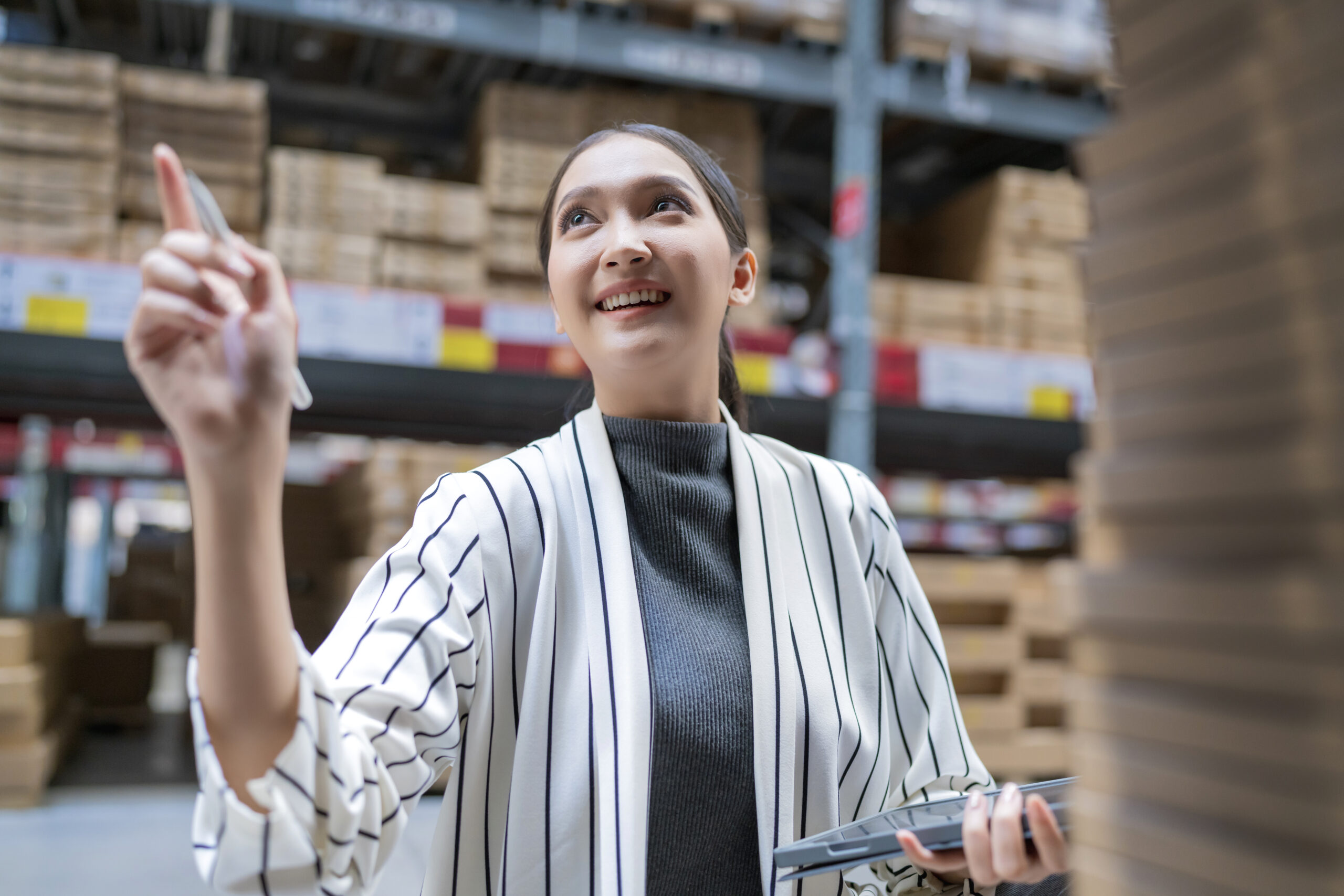 Uma jovem mulher, vestindo um blazer branco com listras verticais pretas e uma blusa cinza de gola alta, sorri enquanto aponta para cima com o dedo indicador da mão direita. Ela segura um tablet preto na mão esquerda e está dentro de um armazém espaçoso, com o fundo preenchido por prateleiras altas de metal estocadas com muitas caixas de papelão.