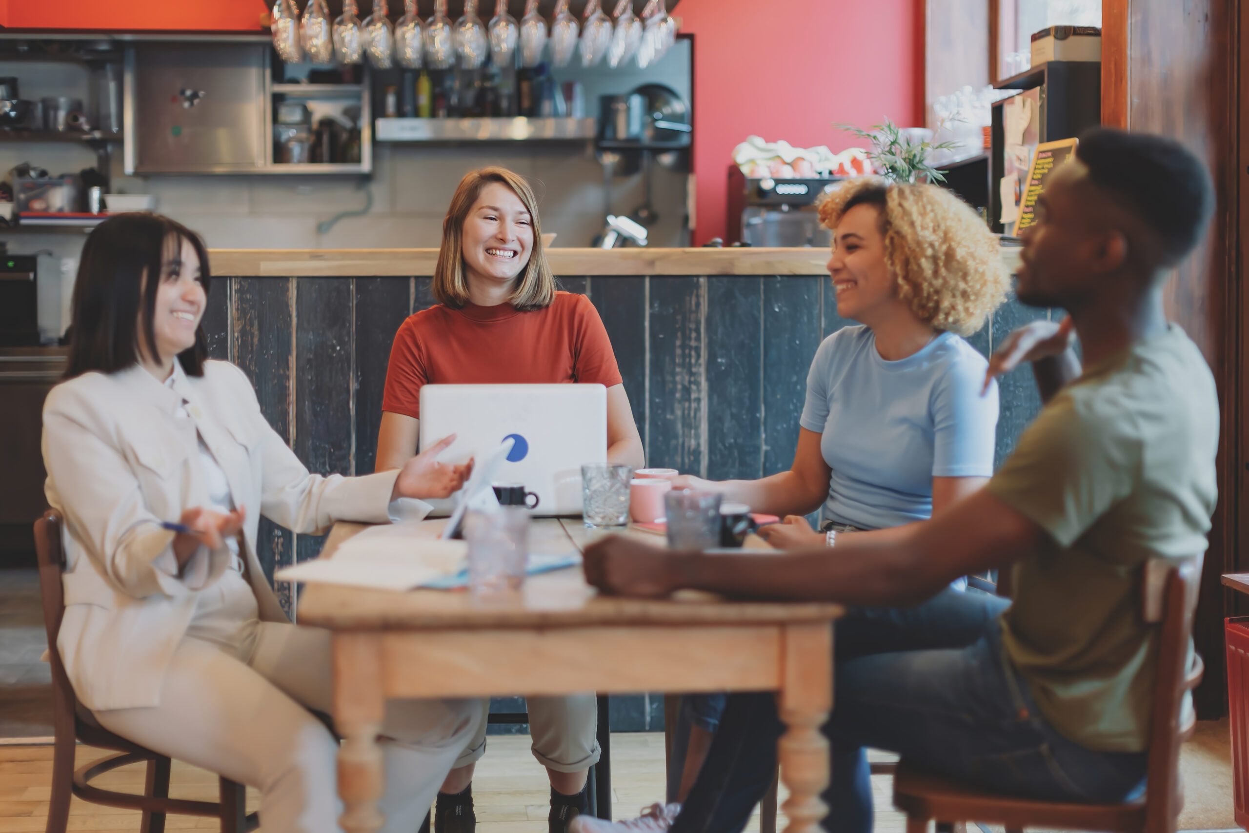 Grupo de pessoas reunido em uma cafeteria, conversando e colaborando em torno de uma mesa com notebooks, cadernos e xícaras, representando trabalho em equipe e produtividade