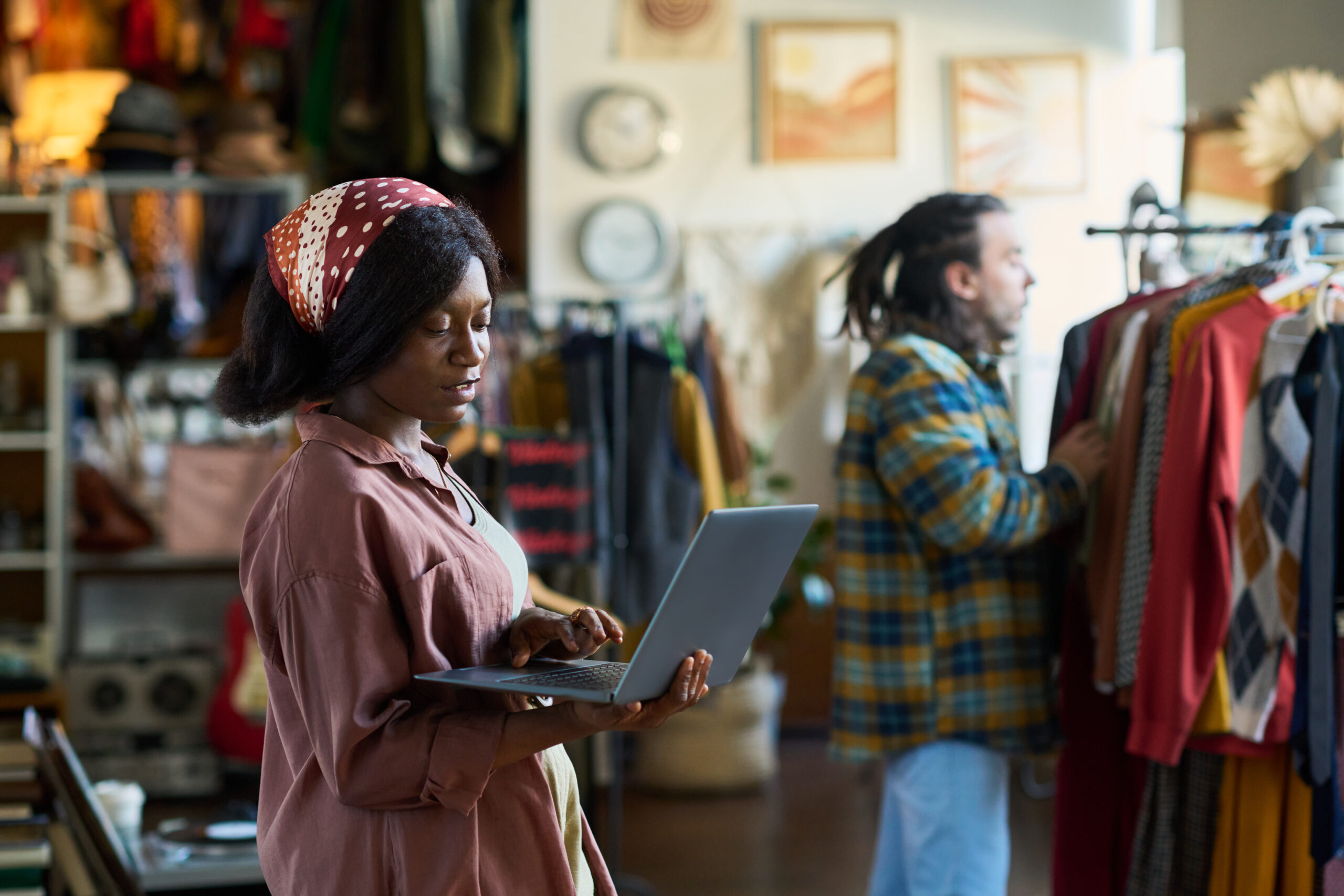 Feature image Profissional usando um notebook dentro de uma loja de varejo enquanto outra pessoa analisa roupas em um cabideiro, representando digitalização, gestão e tendências modernas do varejo.