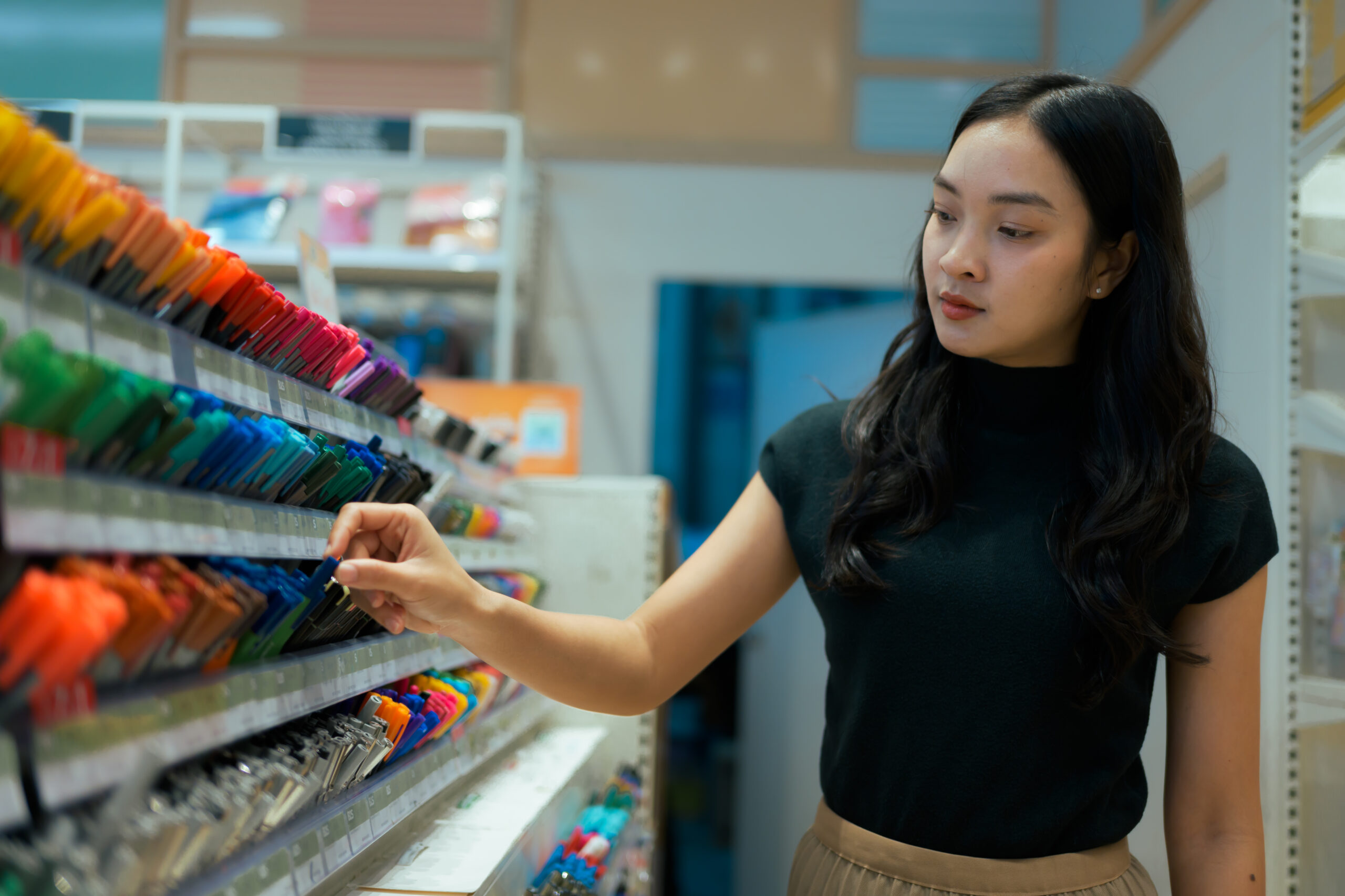 Feature image Mulher asiática escolhendo uma caneta na prateleira dentre várias opções disponíveis na papelaria