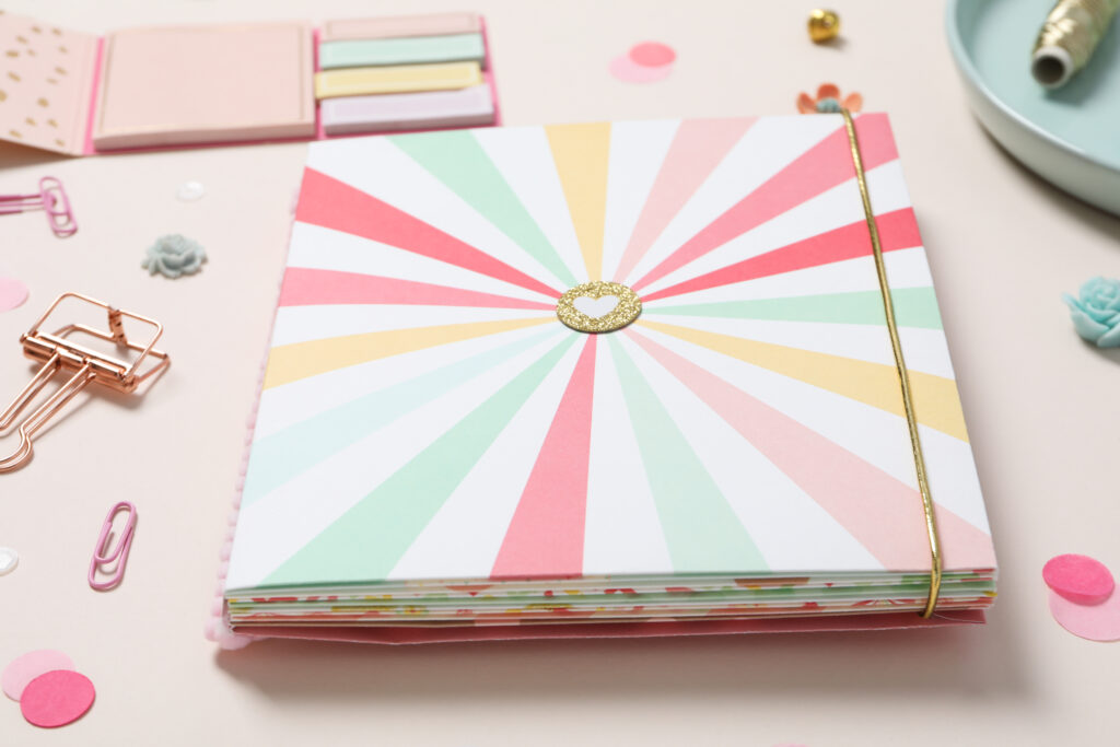 Caderno com estampa colorida, post-its e clips decorativos sobre fundo rosa, exemplos de produtos para vender em papelaria personalizada.