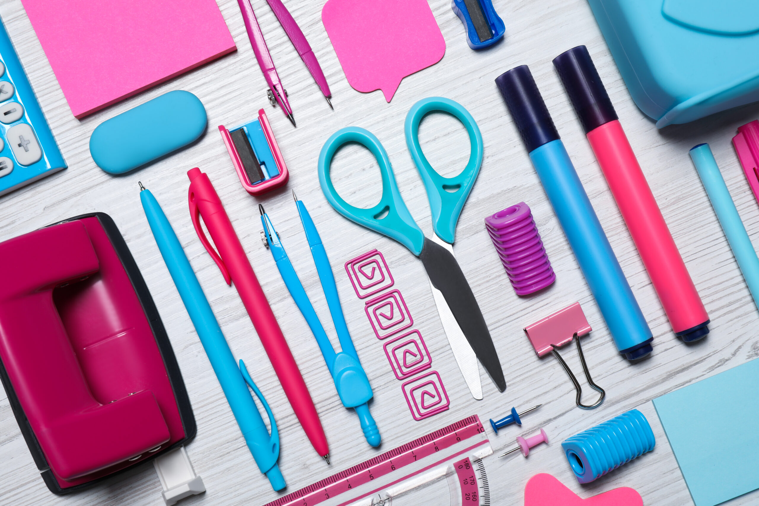 kit de material escolar coloridos, incluindo tesoura, canetas, grampos e furador, organizados sobre superfície branca.